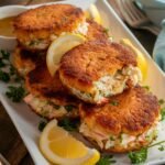 A plate of Gluten Free Crab Cake Recipe with a golden, crispy crust and a tender, flaky interior filled with lump crab meat, fresh herbs, and seasonings. The crab cakes are stacked on a white platter, garnished with parsley and lemon wedges for a fresh, zesty touch. A small dish of dipping sauce sits beside the plate, adding extra flavor. The rustic wooden table and soft natural lighting create a warm, inviting presentation.