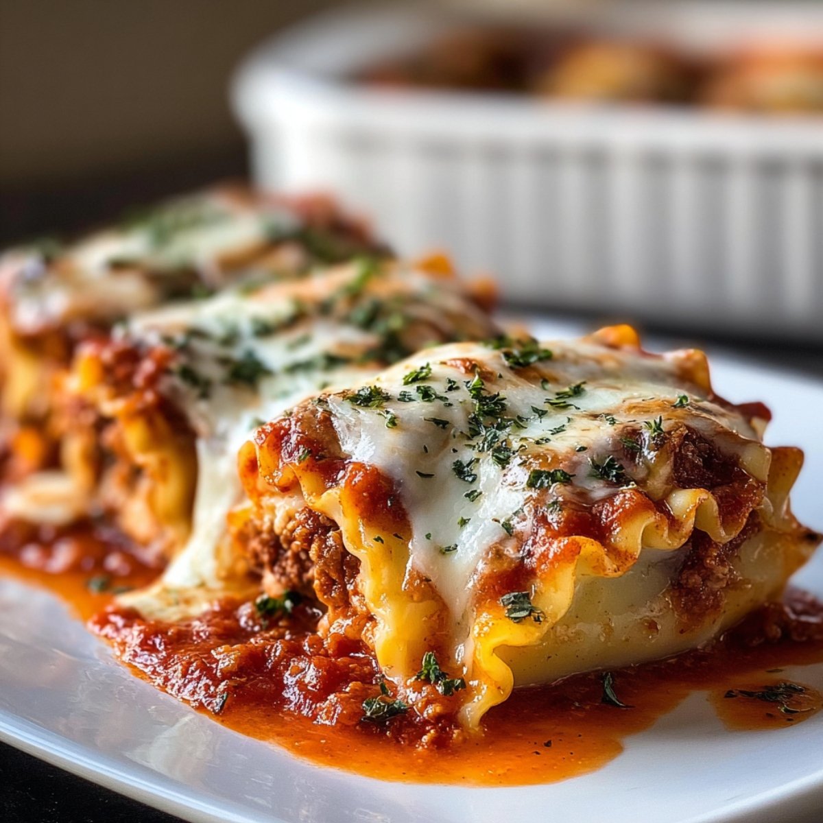 Close-up of homemade lasagna roll ups topped with melted mozzarella and fresh parsley, sitting in rich marinara sauce on a white plate. The pasta is golden and curled, with a meaty ricotta filling visible. A white baking dish with more roll ups is blurred in the background.