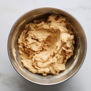 Homemade peanut butter cookie dough in a large stainless steel mixing bowl on a white countertop, showing a soft, creamy texture ready for baking.